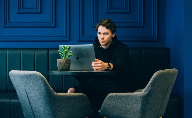 A man sitting against a blue wall at a table with a laptop. He's prepping for an interview question about his biggest weakness.
