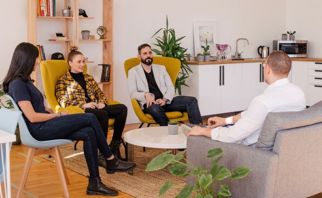 Two women and a man interview another man for a job. They're sitting in comfy chairs in a bright office.
